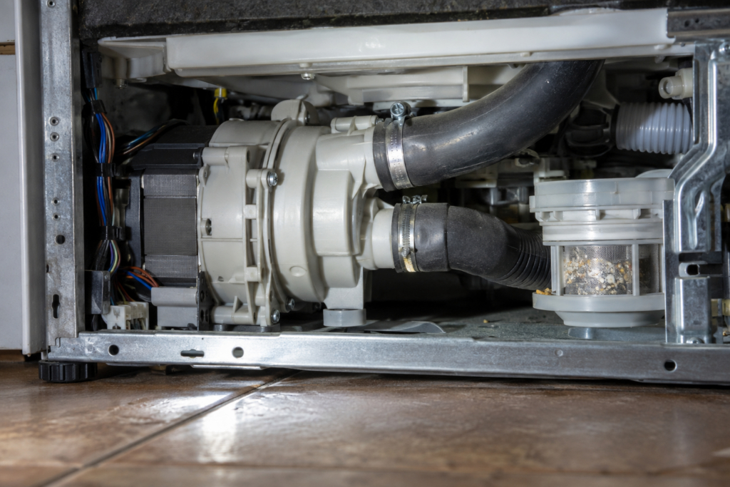 Close-up under a dishwasher with its lower panel removed, showing a plastic drain pump housing with hose connections and a bit of sediment on a nearby filter.