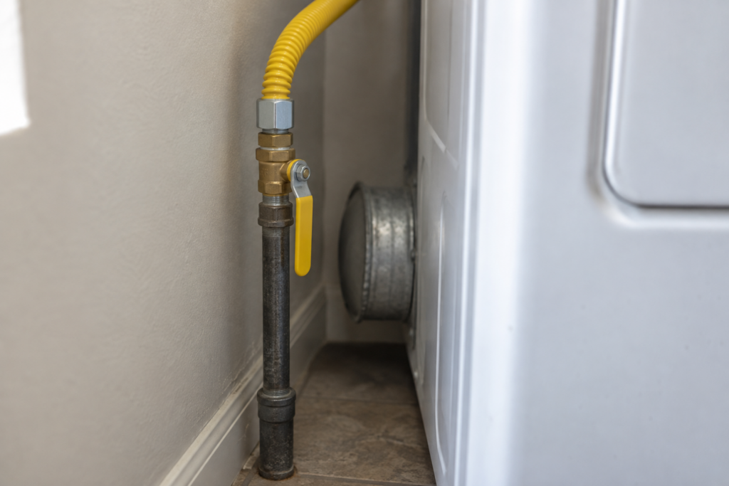 A brass gas shutoff ball valve on the supply line behind a clothes dryer, its lever parallel to the pipe in the open position, a yellow flexible connector attached.