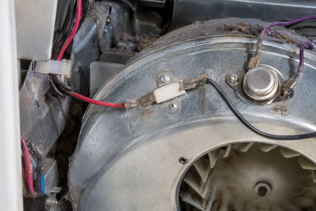 A close view inside the back of a tumble dryer shows a white ceramic thermal fuse and a round metal thermostat on the sheet-metal blower housing under settled lint.