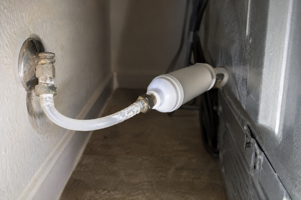 Close-up of a flexible water supply line behind a refrigerator with an inline filter cartridge and fitting, a light chalky mineral residue visible on the tubing.