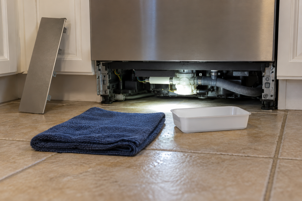A flashlight beam on the floor of the base area beneath a dishwasher with the kick plate removed and leaning against the cabinet, a folded towel and a small plastic tray on the tiles in front.