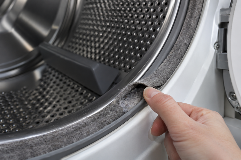 A fingertip lifts a frayed section of the felt seal along the front edge of a dryer drum.