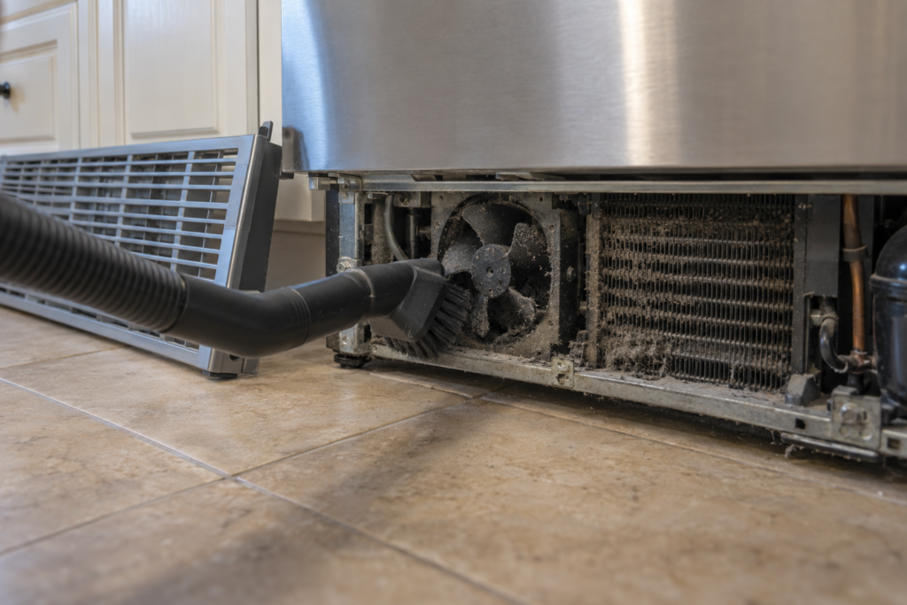 The base grille of a refrigerator removed and leaning on the cabinet, a vacuum brush attachment reaching toward a small condenser fan and coil coated in grey dust beneath the cabinet, side light.