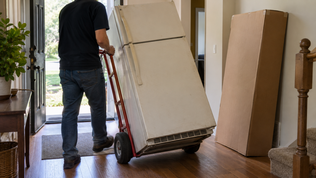 An old refrigerator on a hand truck near an open front door with a new appliance box beside it.